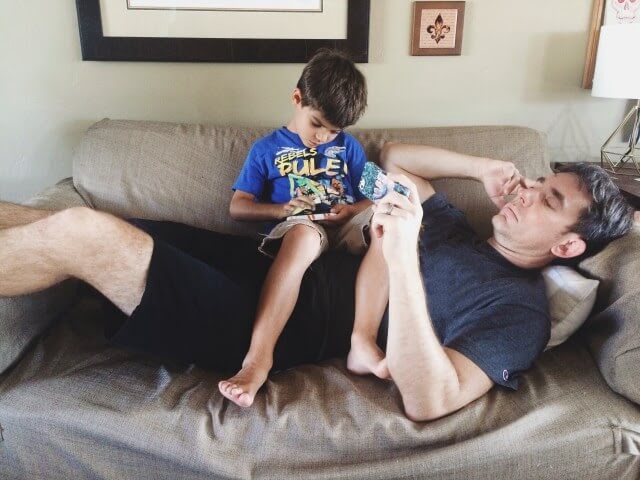A kid playing with gadget sitting on father's lap , meanwhile father is also busy on his phone
